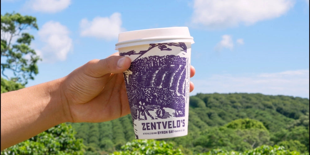 Hand holding a Zentvelo's coffee cup with a scenic background of trees and blue sky.